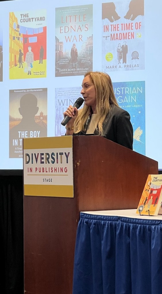 Janet Bond Brill speaking at the Diversity in Publishing stage, with Little Edna’s War projected behind her.